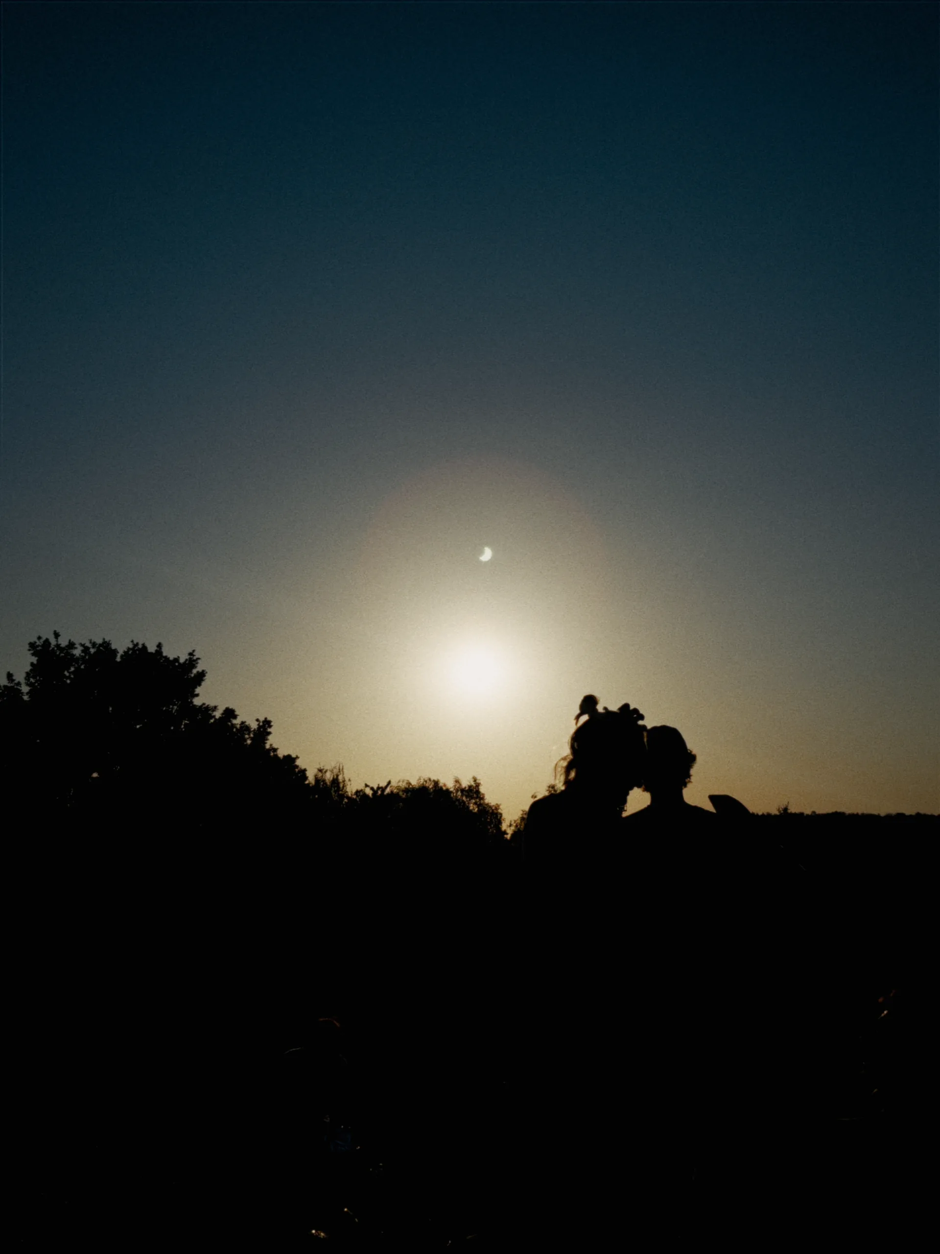 Two people silhouetted against a glowing sky, with a thin crescent hanging above them.