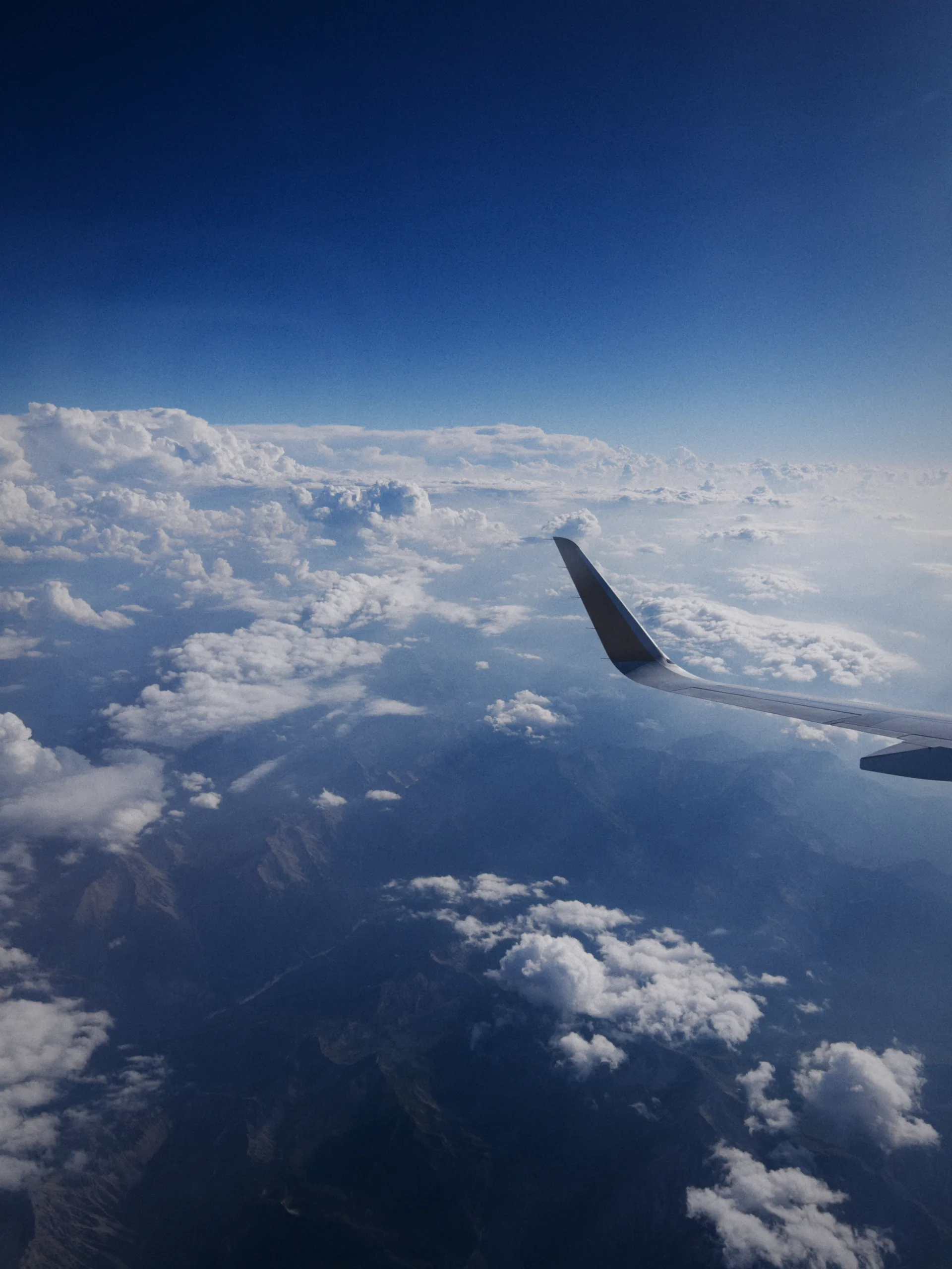 An aircraft wing above a layer of cloud, with mountains far below and deep blue sky above.